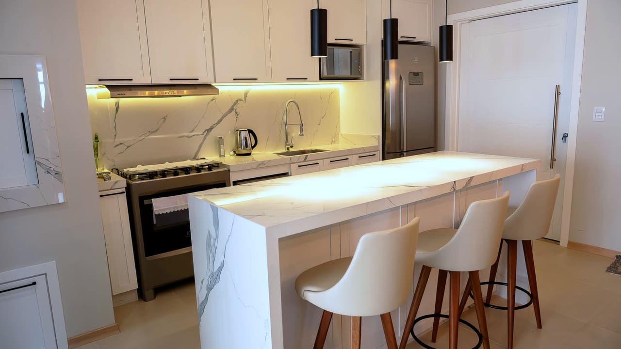Cozinha com ilha de mármore branco, pendentes pretos, banquetas de madeira e iluminação embutida sob os armários