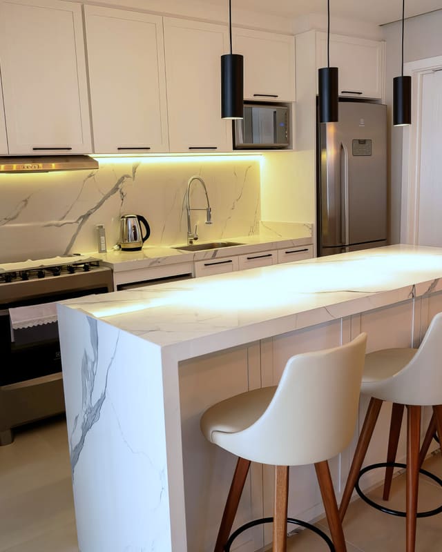 Cozinha com ilha de mármore branco, pendentes pretos, banquetas de madeira e iluminação embutida sob os armários