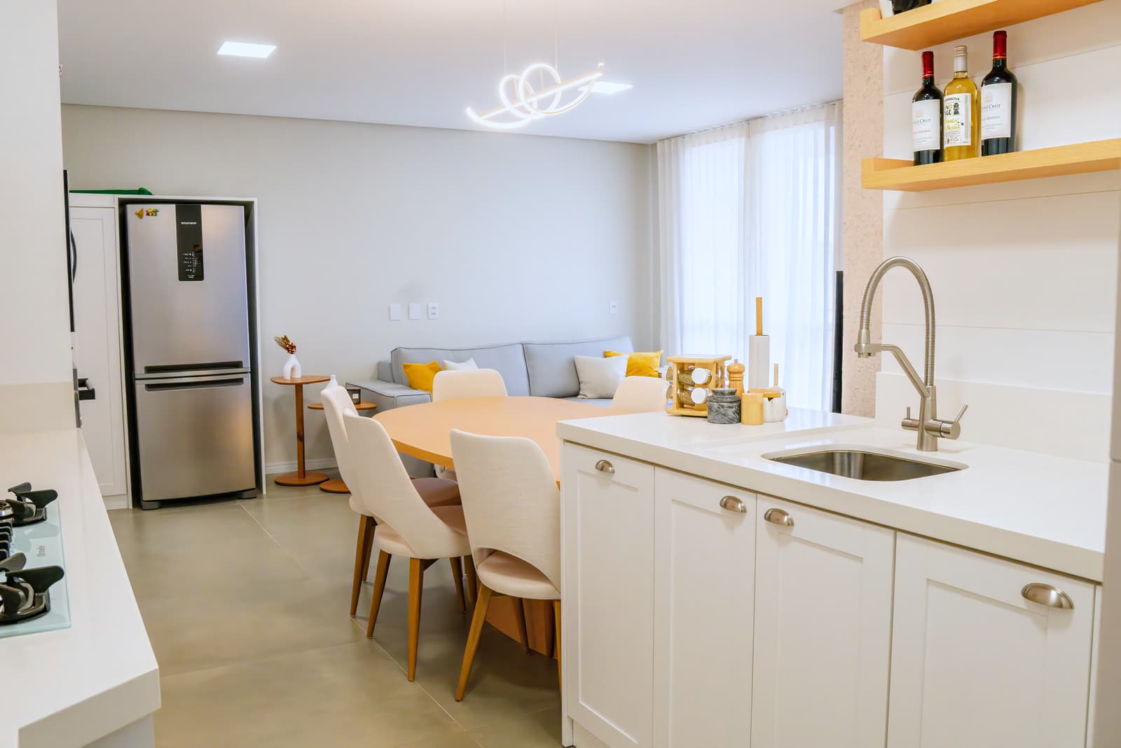 Cozinha e sala de jantar integradas, com marcenaria branca, prateleiras de madeira clara e mesa oval
