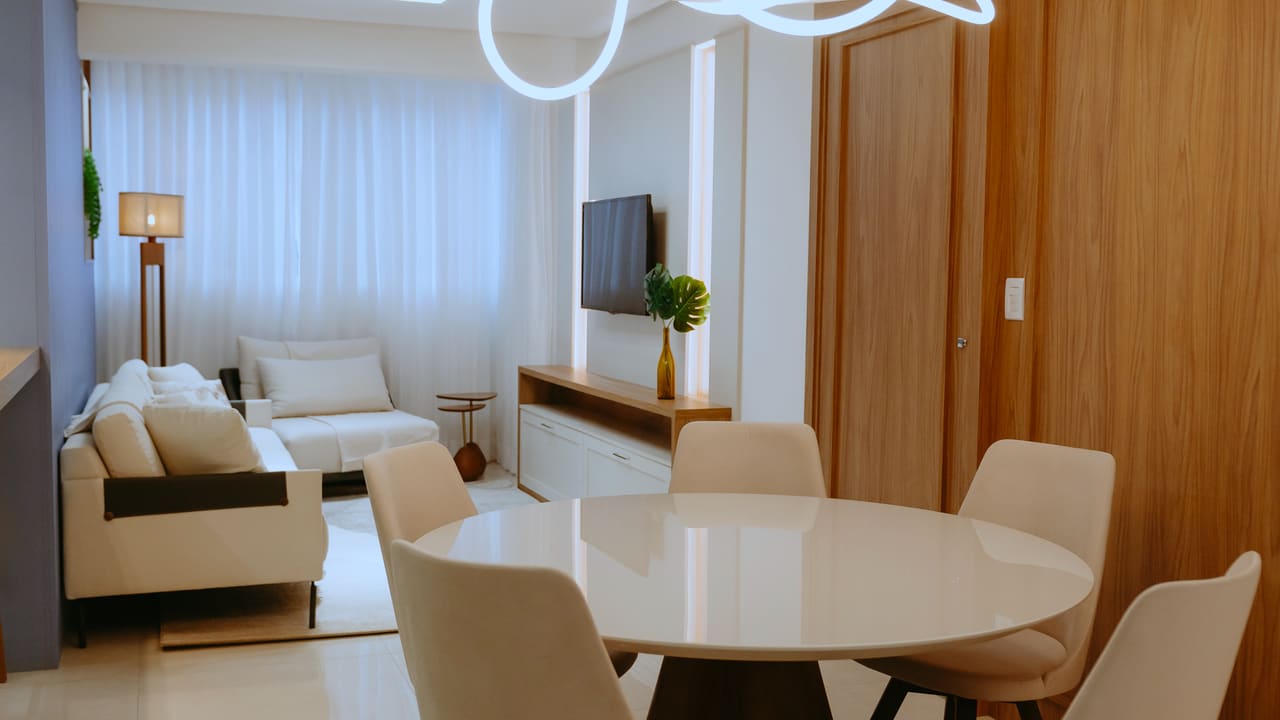Sala de estar e jantar integradas, com sofá claro, mesa redonda, marcenaria de madeira e luminária de neon contínuo no teto