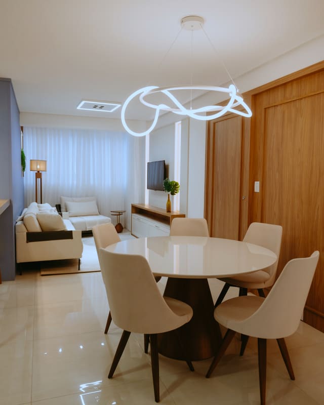 Sala de estar e jantar integradas, com sofá claro, mesa redonda, marcenaria de madeira e luminária de neon contínuo no teto