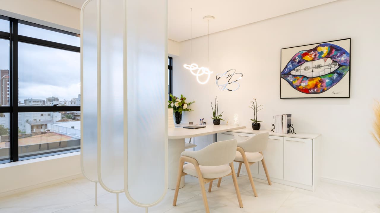 Sala de consultório odontológico com divisórias curvas em vidro leitoso, mesa branca, cadeiras de madeira clara e luminária de neon
