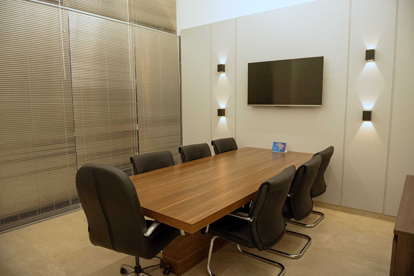 Sala de reunião com mesa de madeira, cadeiras escuras e arandelas nas duas faces