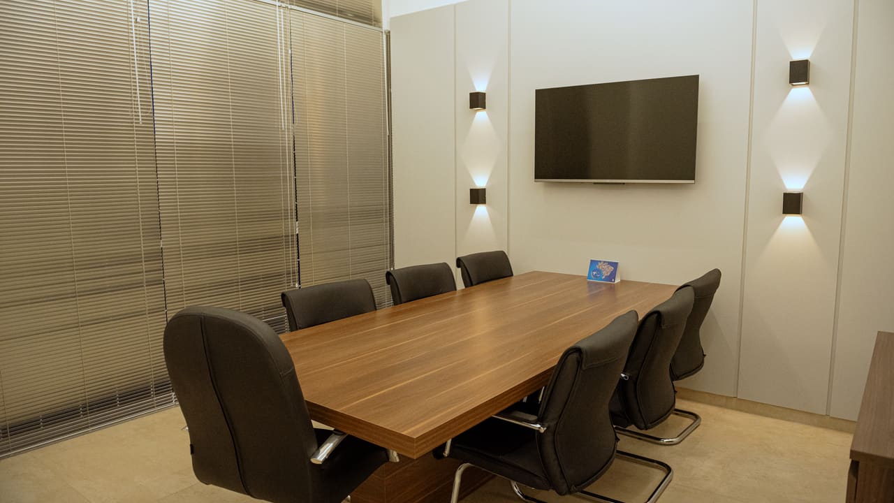 Sala de reunião com mesa de madeira, cadeiras escuras e arandelas de luz indireta nas duas faces da parede
