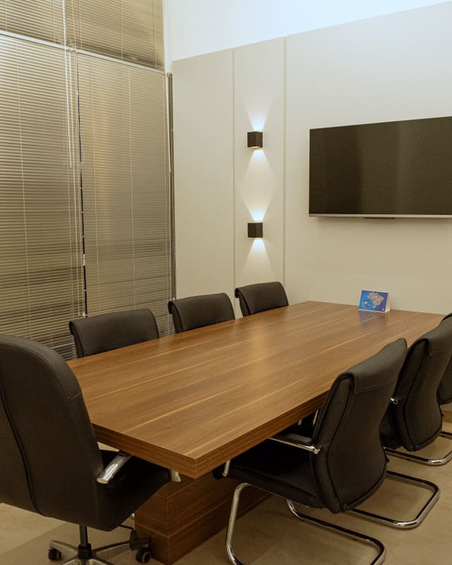 Sala de reunião com mesa de madeira, cadeiras escuras e arandelas de luz indireta nas duas faces da parede