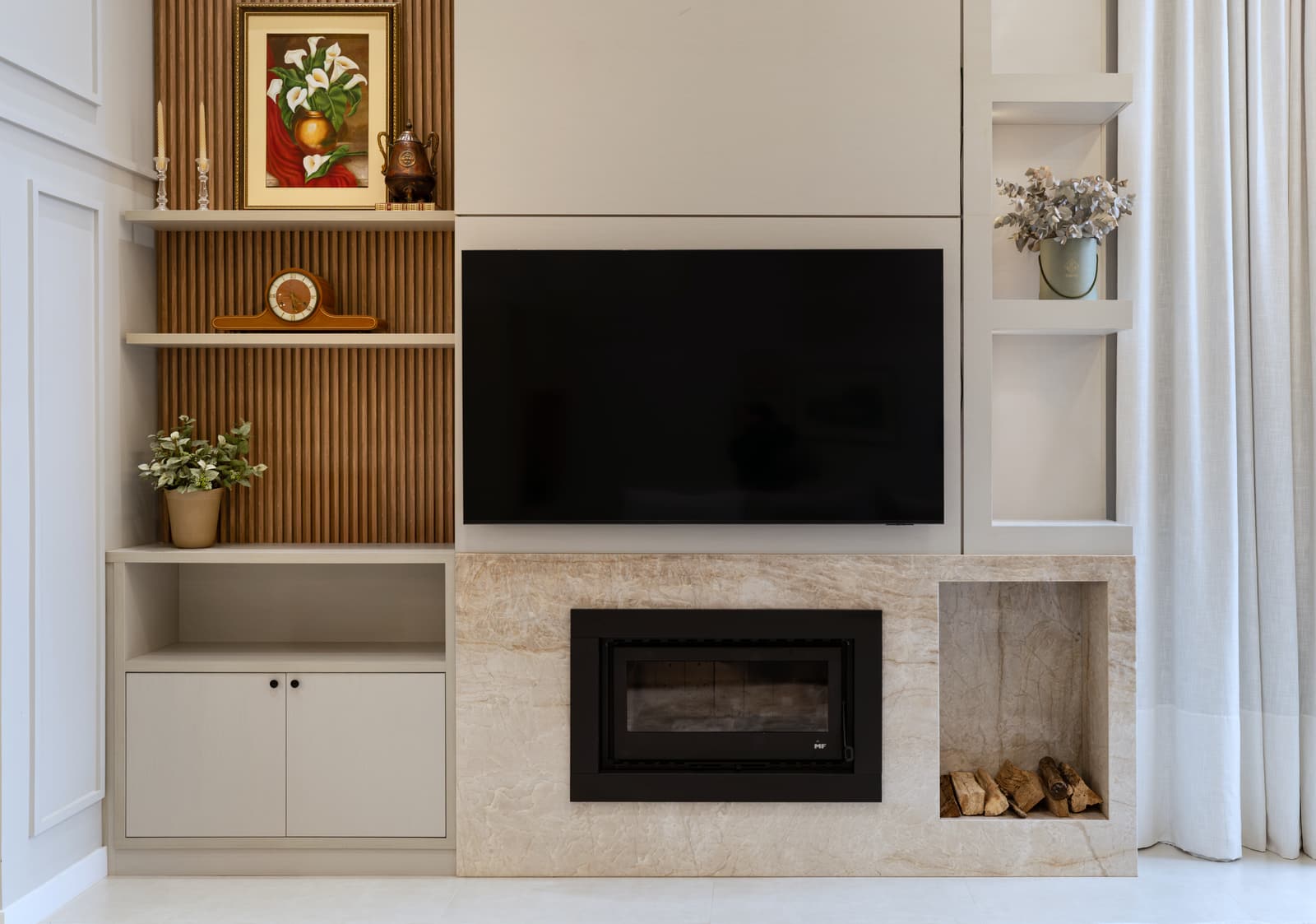 Parede da sala de TV, com lareira em mármore, boiserie clara e nicho de lenha