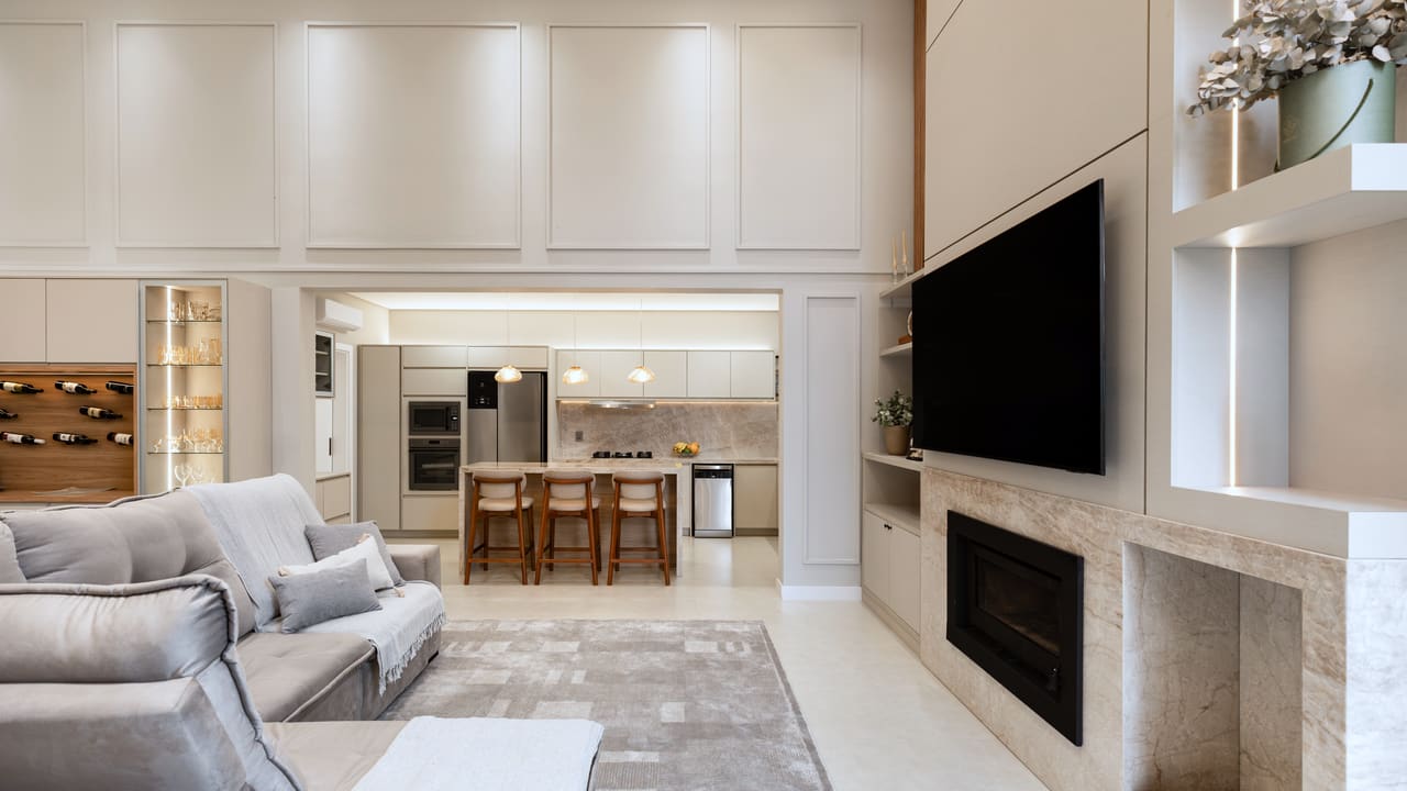 Sala de estar com boiserie clara, lareira revestida em mármore e vista para a cozinha com ilha e banquetas de madeira