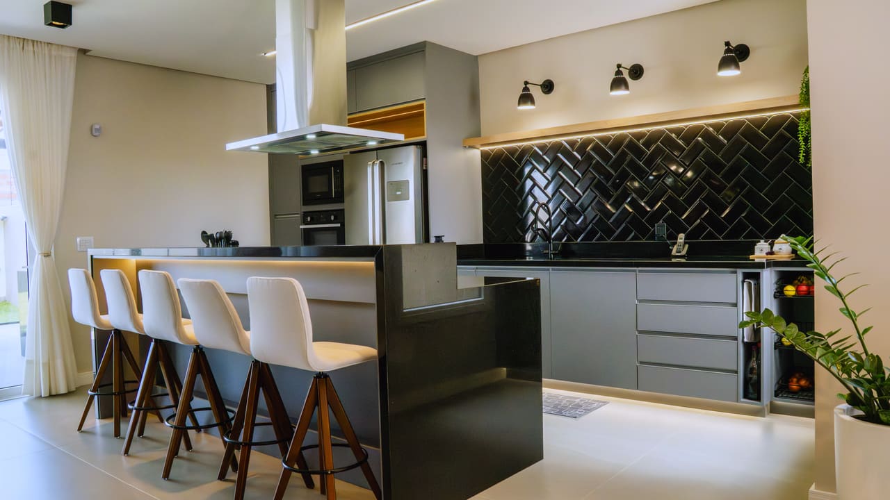 Cozinha com ilha central, bancada preta, revestimento em espinha de peixe escuro e banquetas de madeira e estofado claro