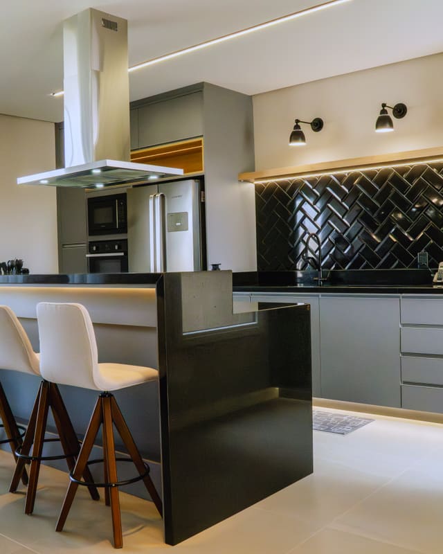Cozinha com ilha central, bancada preta, revestimento em espinha de peixe escuro e banquetas de madeira e estofado claro
