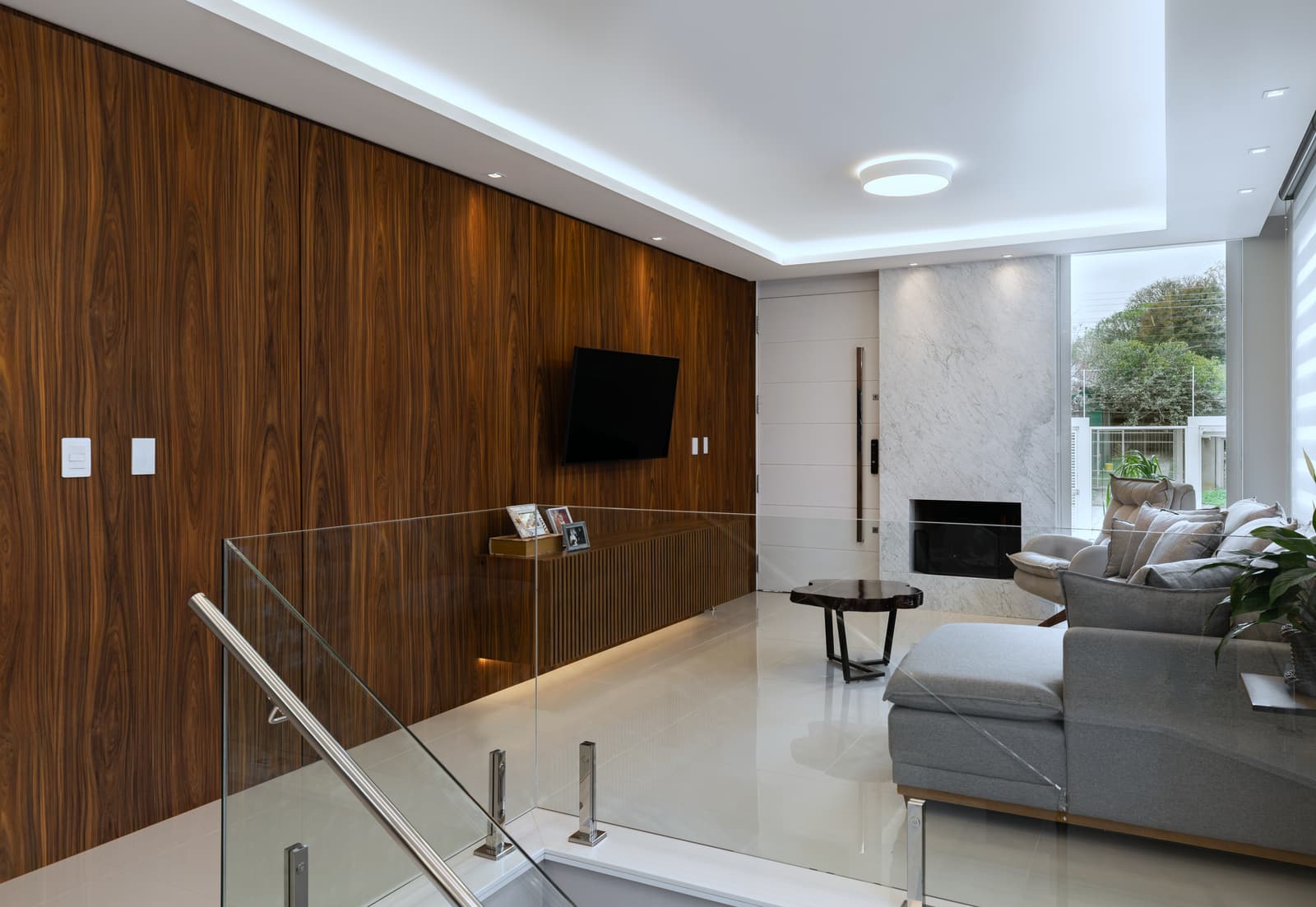 Hall do piso superior com guarda-corpo de vidro, painel de madeira e vista para o estar com lareira