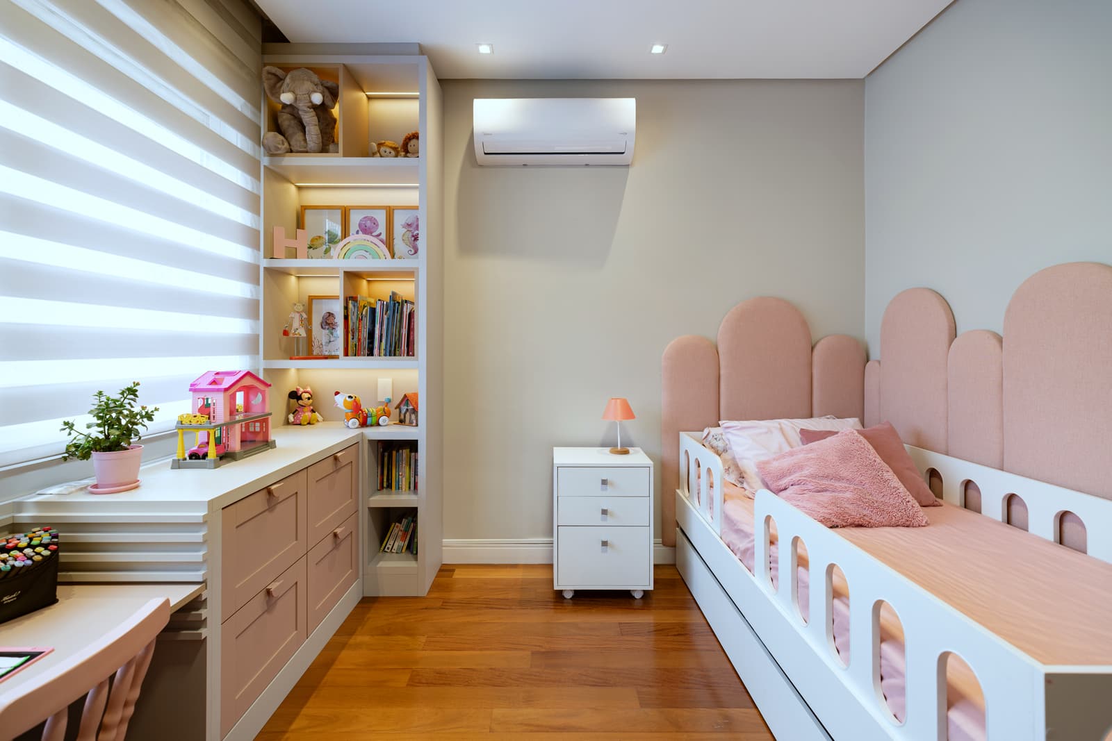 Quarto infantil com cabeceira em arcos rosa, escrivaninha sob a janela e estante iluminada