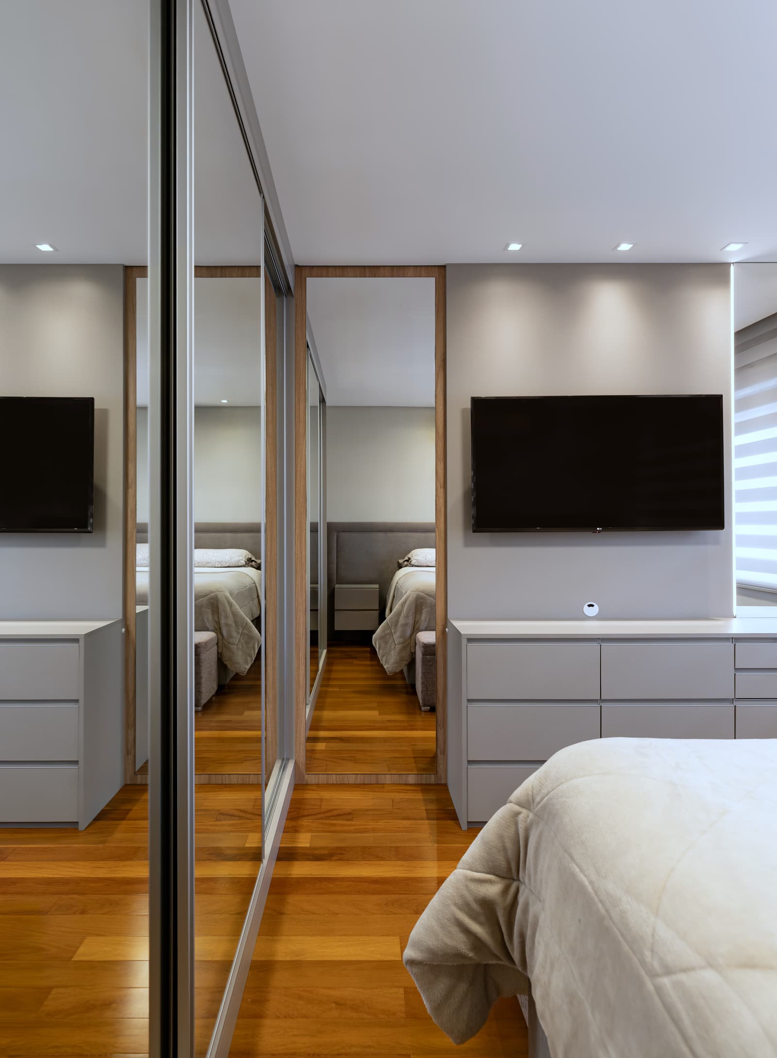 Suíte do casal vista da cama, com televisão na parede e espelho de correr refletindo o quarto