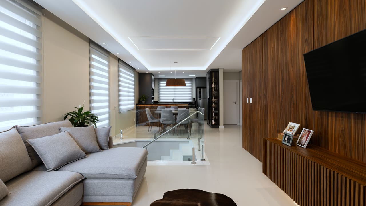 Sala de estar no piso superior, com sofá cinza, painel de madeira ripada na parede da TV e guarda-corpo de vidro