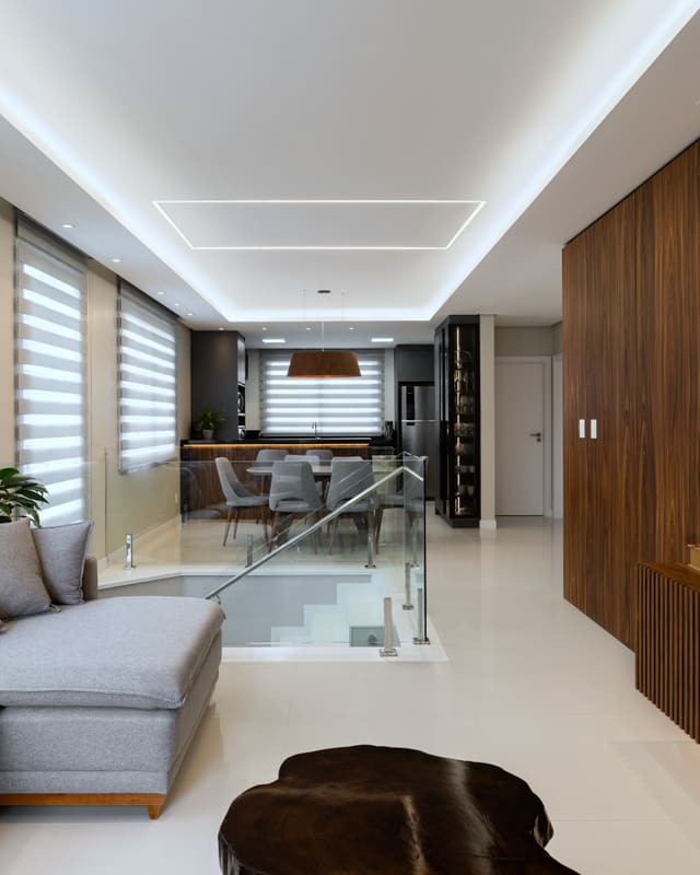 Sala de estar no piso superior, com sofá cinza, painel de madeira ripada na parede da TV e guarda-corpo de vidro